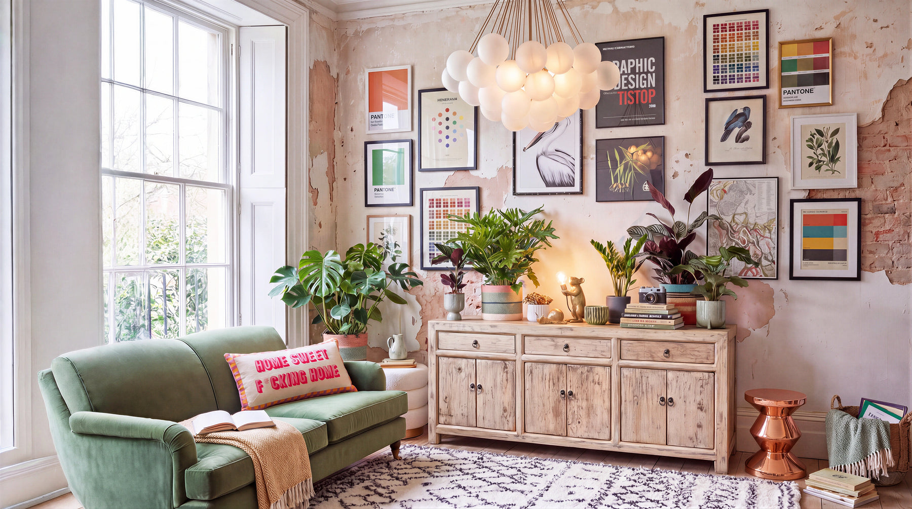 A styled interior room scene with a green velvet sofa, a light wood sideboard, many framed art prints on the wall, potted plants, a hanging cloud-shaped pendant light, and a patterned rug.