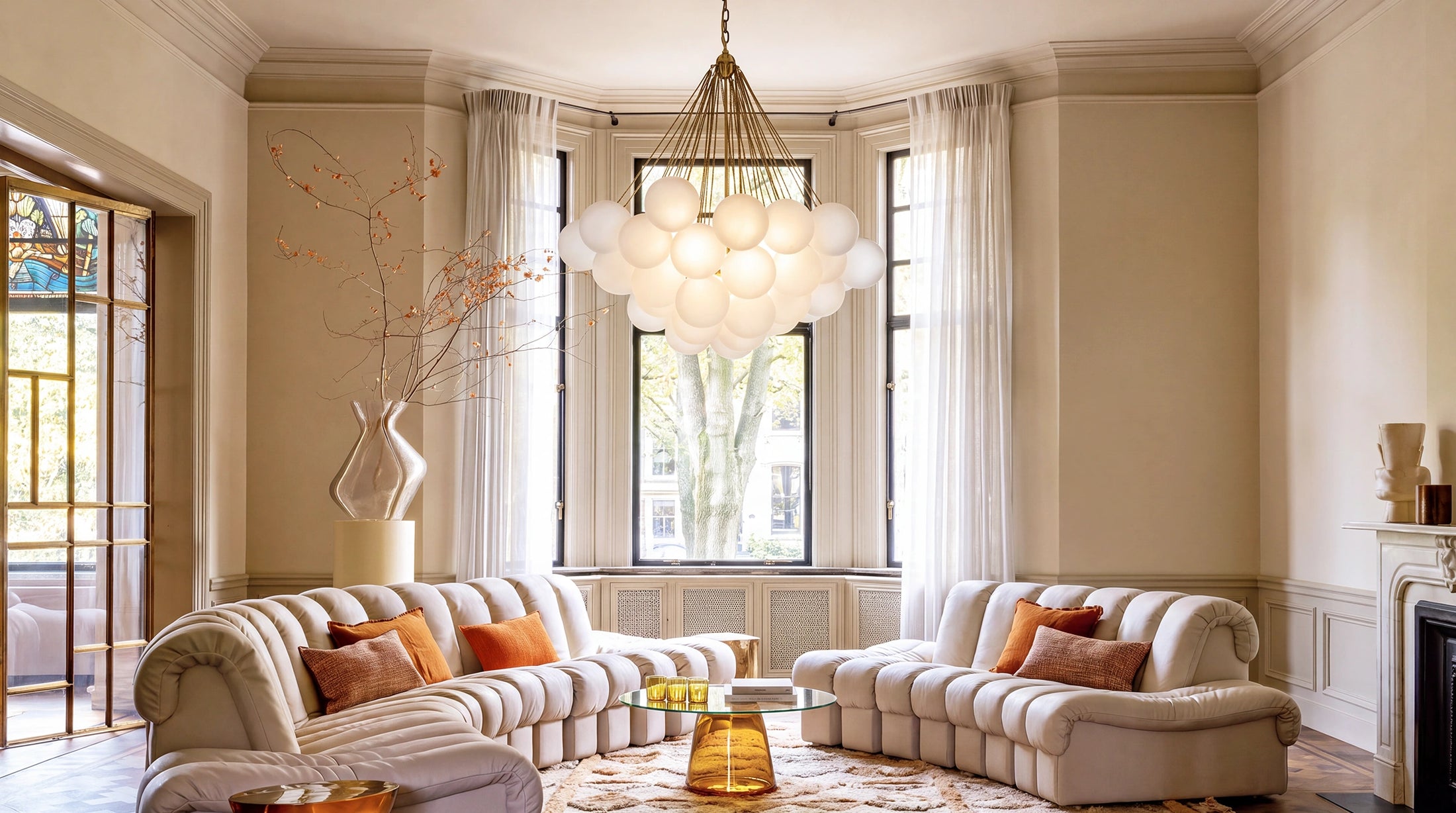 A luxury cloud-shaped chandelier with glowing white glass globes hangs in a bright, elegant living room with cream tufted sofas, sheer curtains, and a bay window.