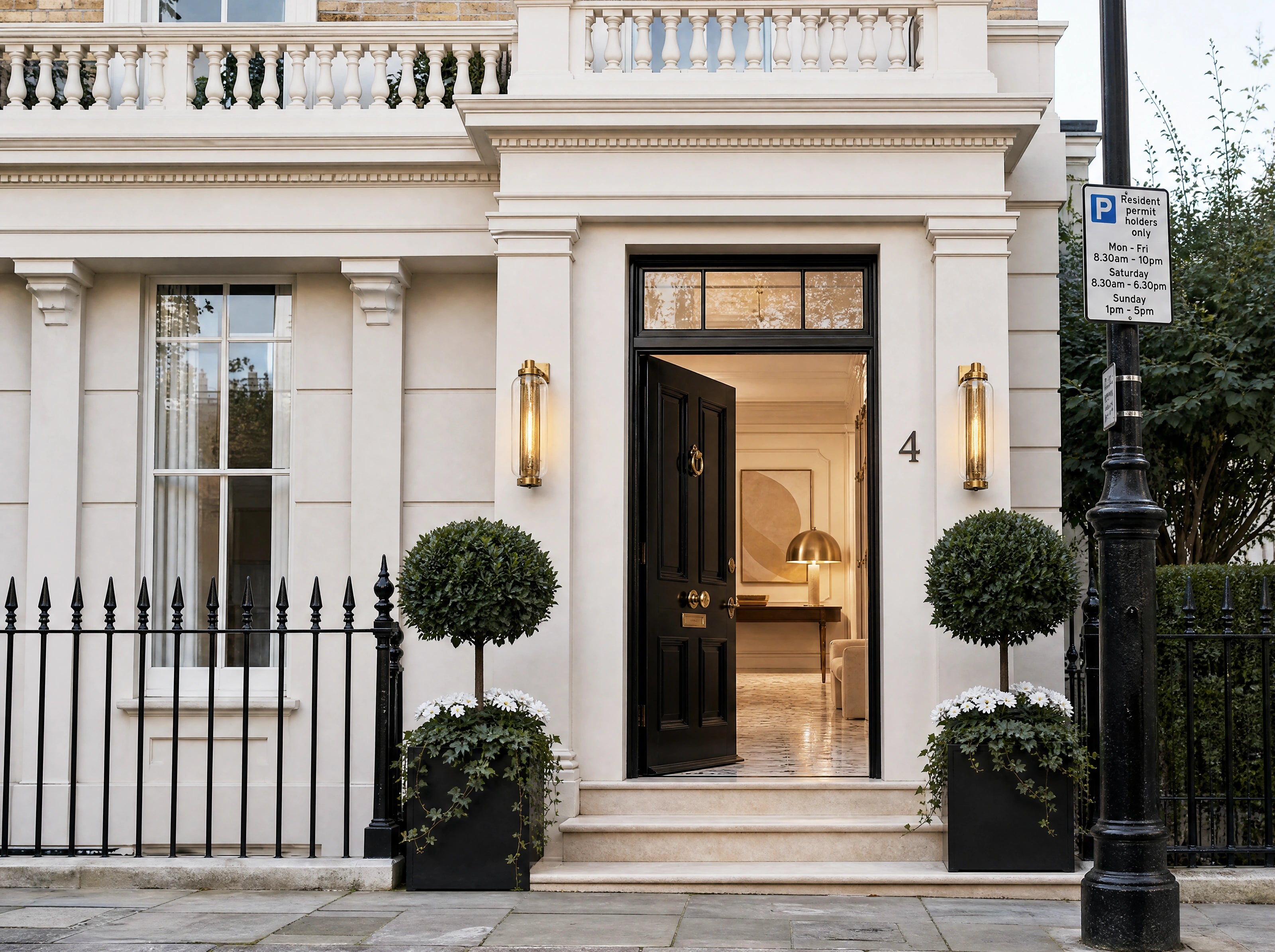 A grand white stone building facade with an open black front door, black iron fence, manicured topiary trees, and a black street lamp on the right.